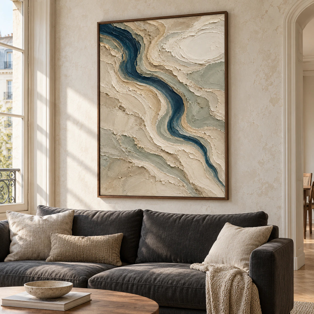 Large vertical painting above a low charcoal sofa in a bright living room, its winding blue-green channel and textured neutral ridges clearly visible.