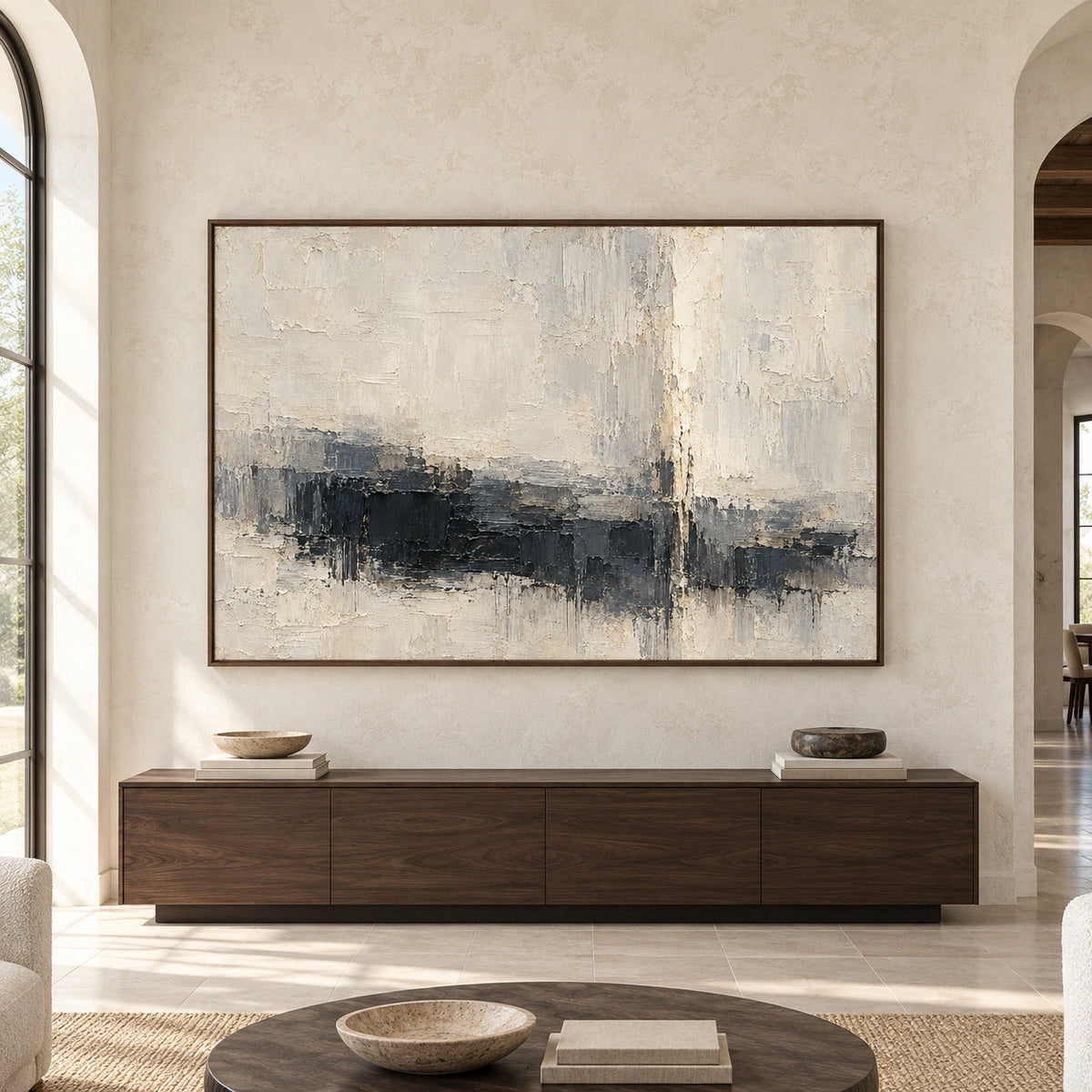 Straight-on view of a large charcoal and ivory abstract painting on a pale stone wall above a low dark wood console in a spacious, daylit room.