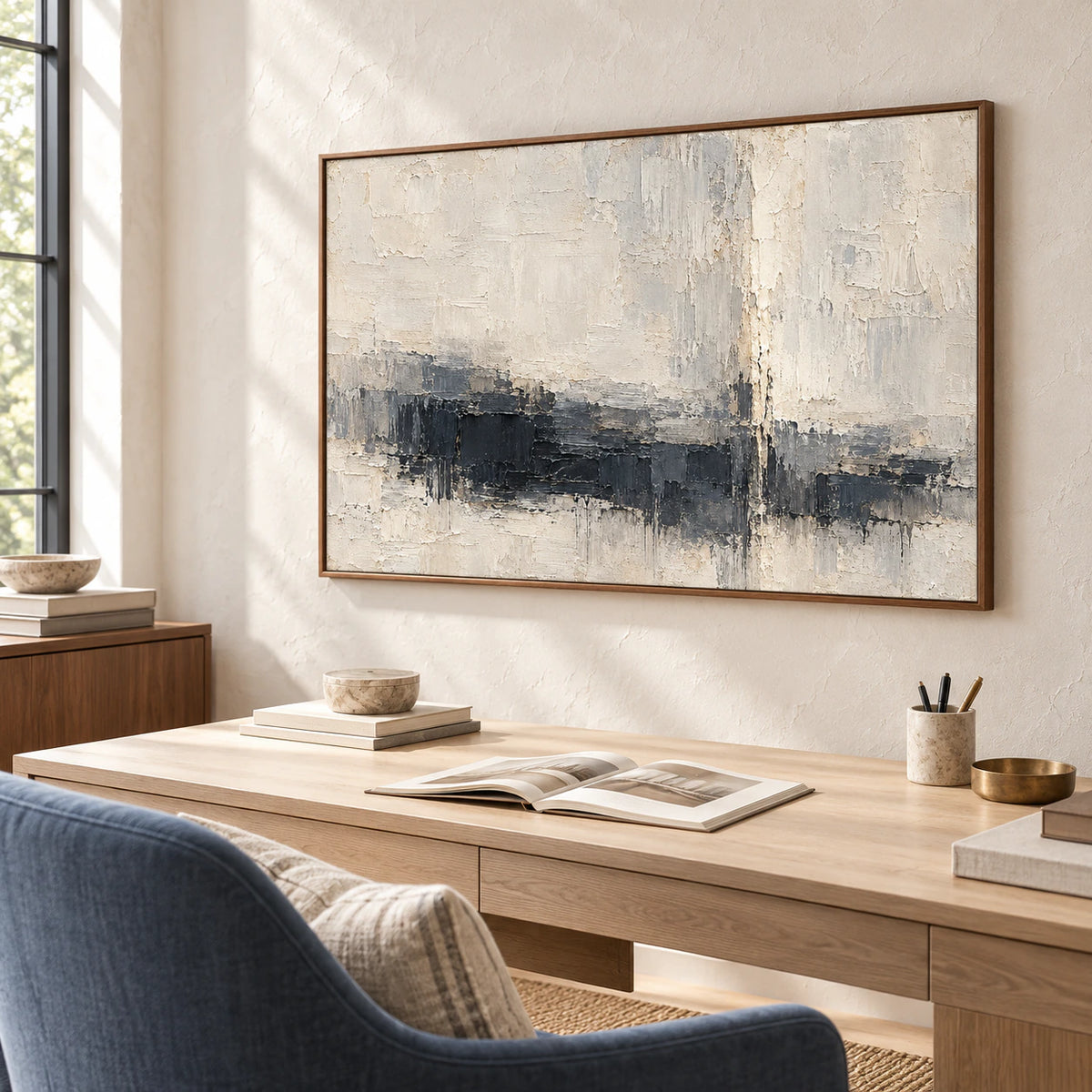 Charcoal and ivory abstract painting above a pale wood desk with a muted blue chair in an airy home office, lit by a tall window and viewed from a higher right angle.