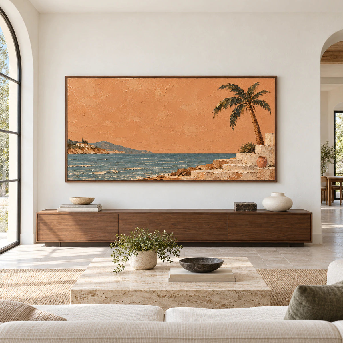 Large framed view of Copper-Littoral above a low console in a spacious, bright room, showing the terracotta sky, blue-green sea, distant village, and palm at impressive scale.
