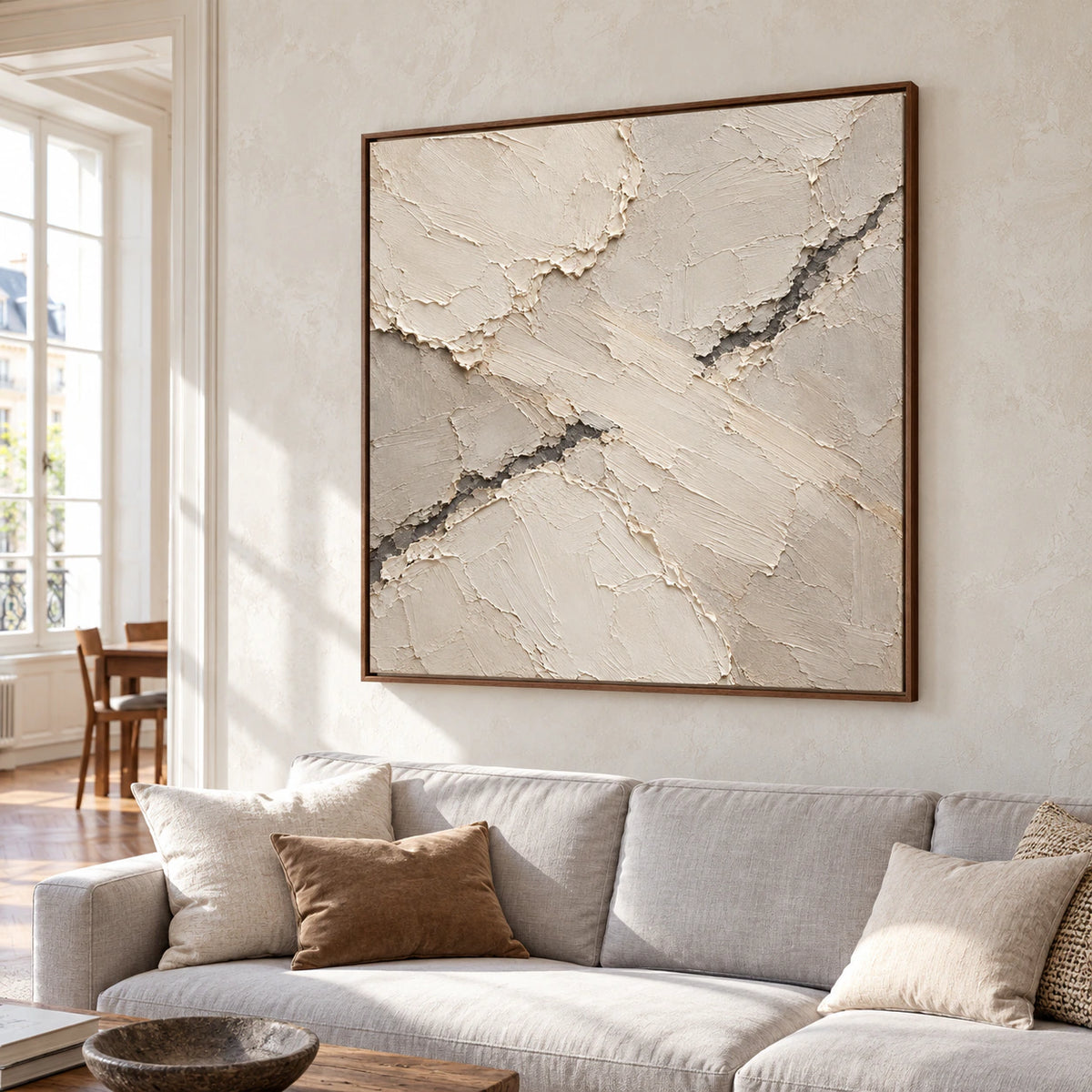 Large square textured artwork above a restrained pale sofa in a bright living room, its diagonal ivory and taupe planes distinct against a soft gray wall.