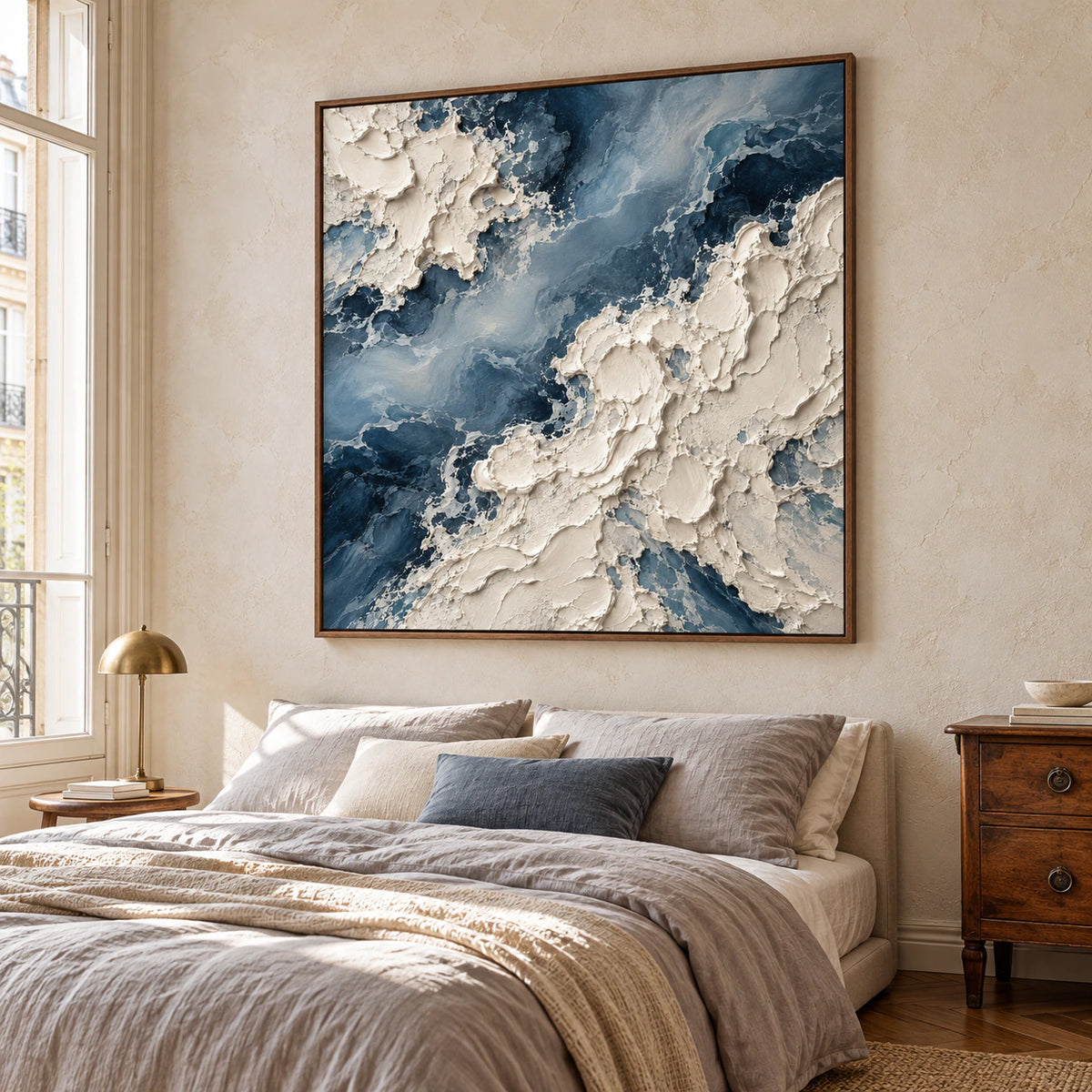 Square blue-and-ivory impasto painting above a low bed with soft gray linen and warm wood details, viewed from the left near bed height in daylight.