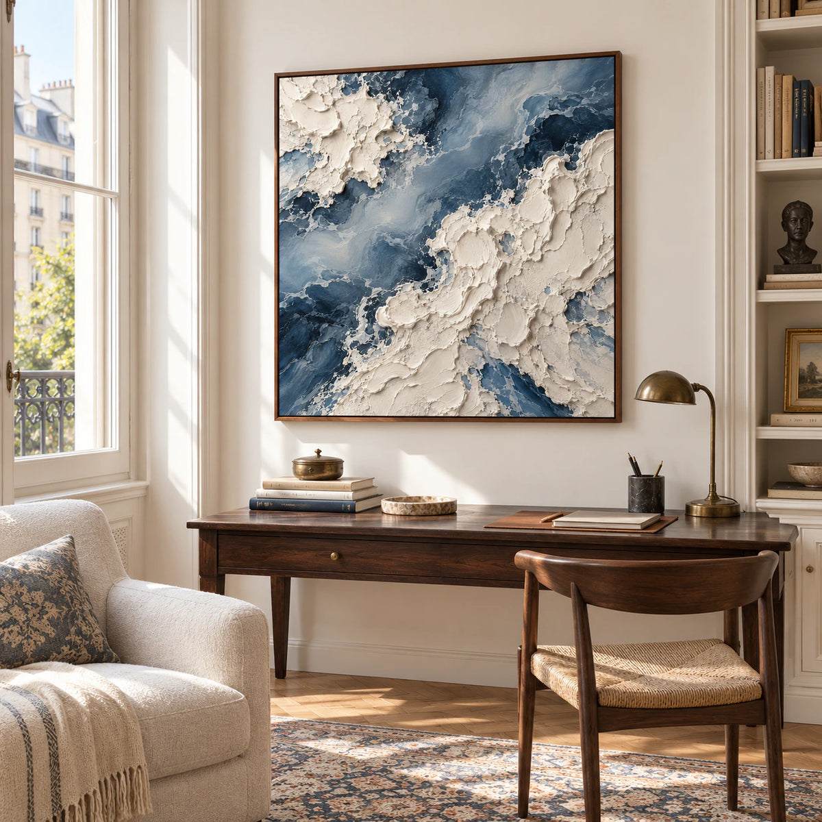 Square blue-and-ivory abstract painting above a dark timber desk beside a pale upholstered reading chair in a bright home office, viewed from the right.