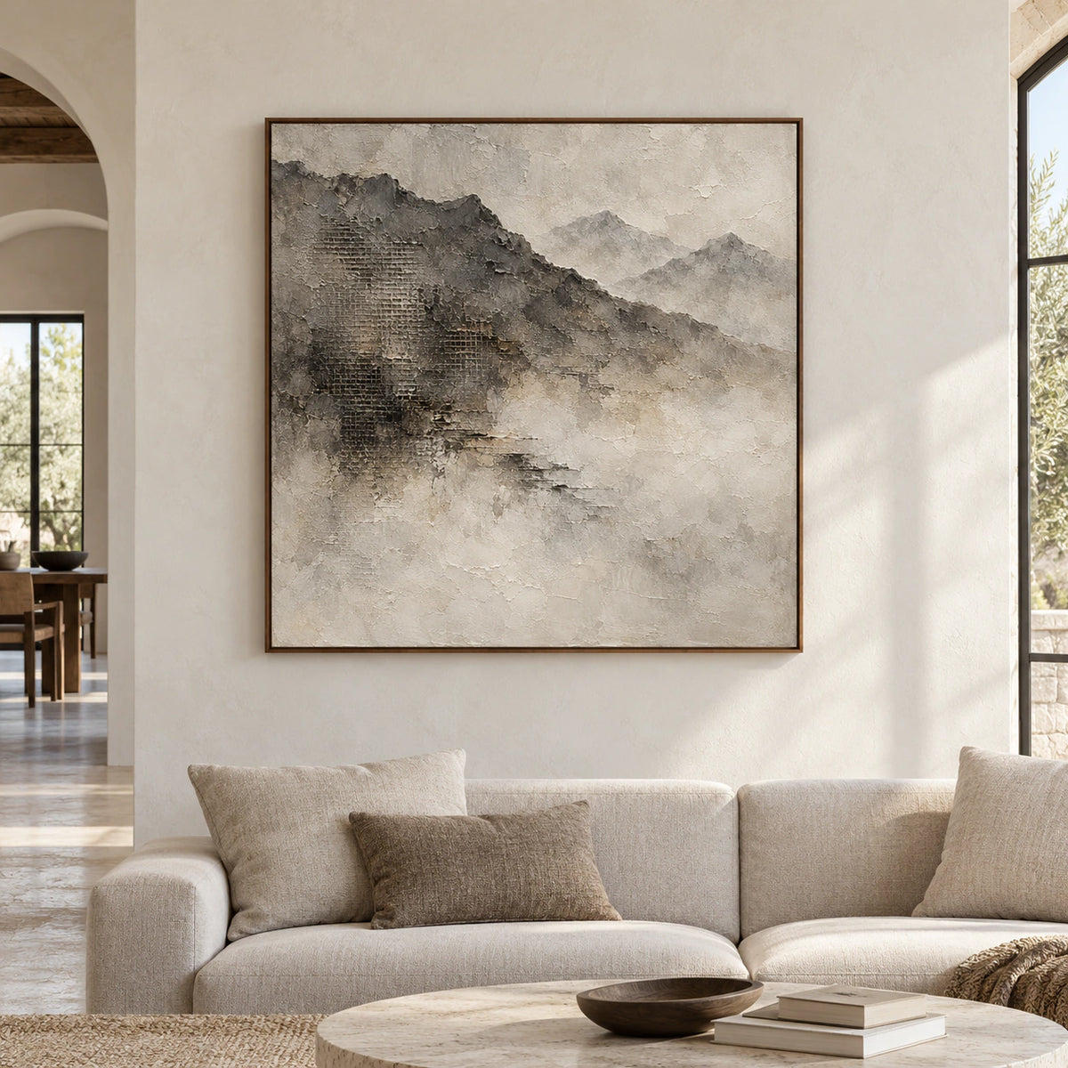 Square mountain painting in a slim brown frame on a broad pale plaster wall in a spacious contemporary room, viewed straight on in natural daylight.