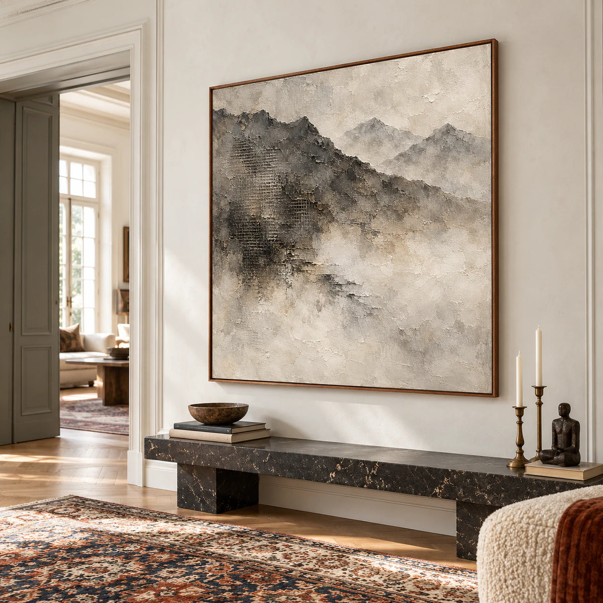 Square mountain painting on a tall light plaster wall above a low dark stone bench in a spacious foyer with a visible doorway, viewed from a low right angle.