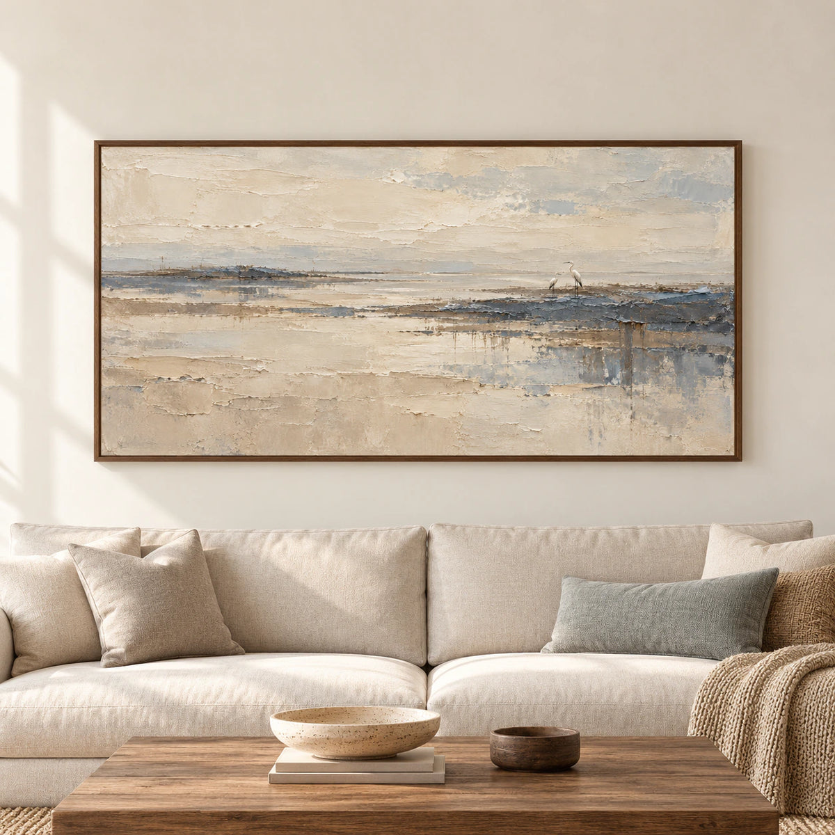 Large horizontal heron shoreline painting above a cream sofa against a warm pale wall in a spacious, daylit interior, viewed straight on.