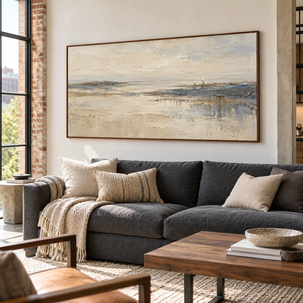 Large heron shoreline painting above a charcoal sofa in a bright living room with tall windows and natural furnishings, viewed slightly from the left.