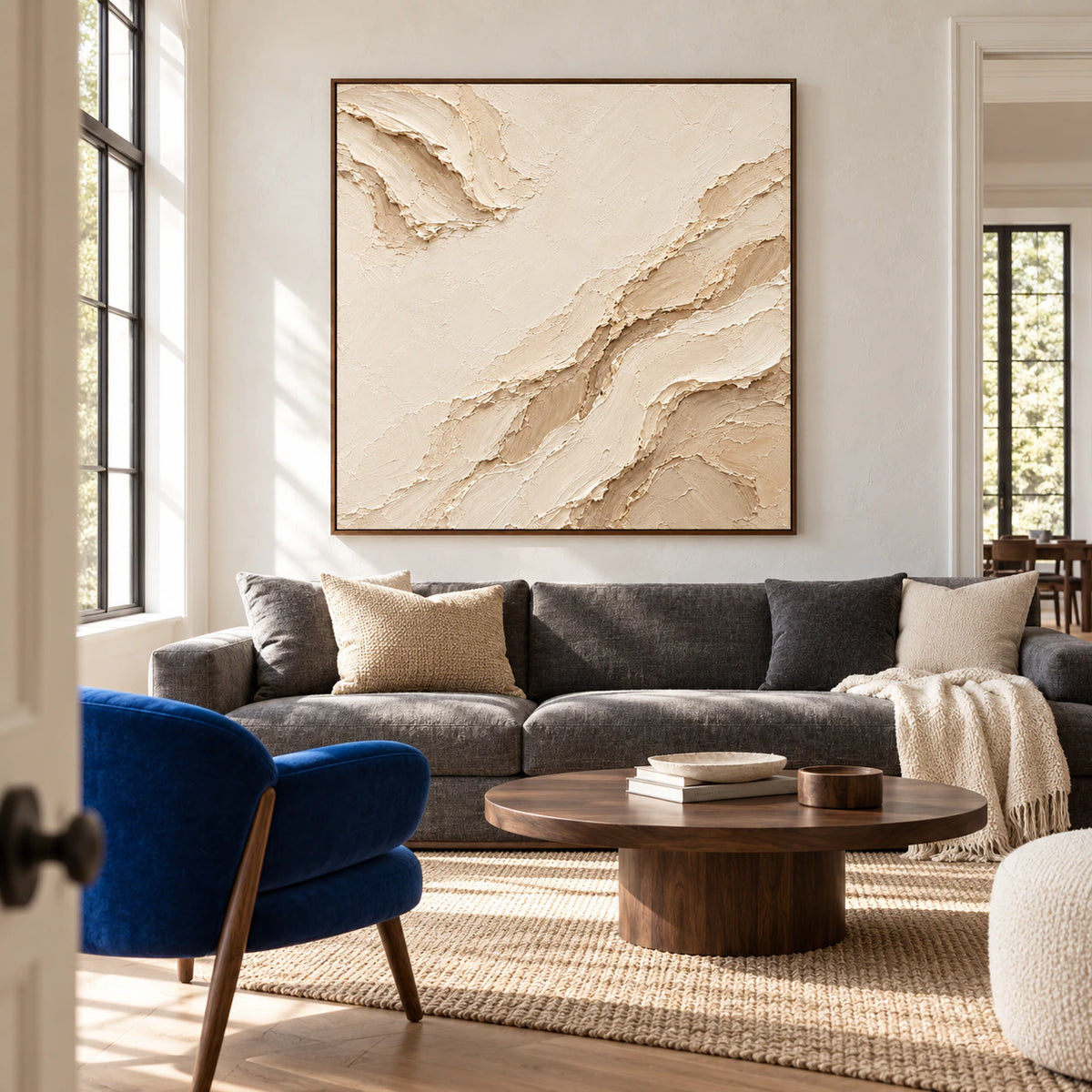 Square ivory and sand abstract painting above a charcoal grey sofa, with broad windows and a dark wood coffee table, viewed from the left.