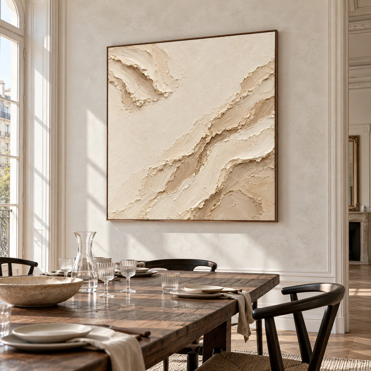 Square ivory and sand abstract painting on a pale stone wall beside a dark oak dining table, black chairs and glassware, viewed from the right.