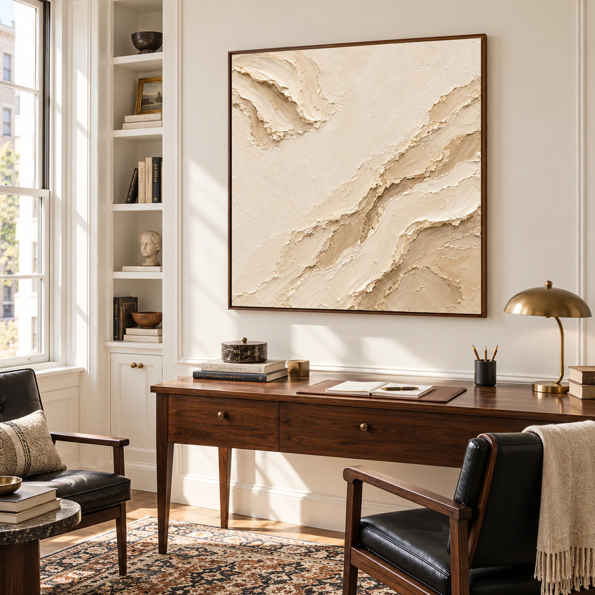 Square ivory and sand abstract painting above a dark walnut wood desk, with a black reading chair, open shelving and a tall window, viewed from the right.