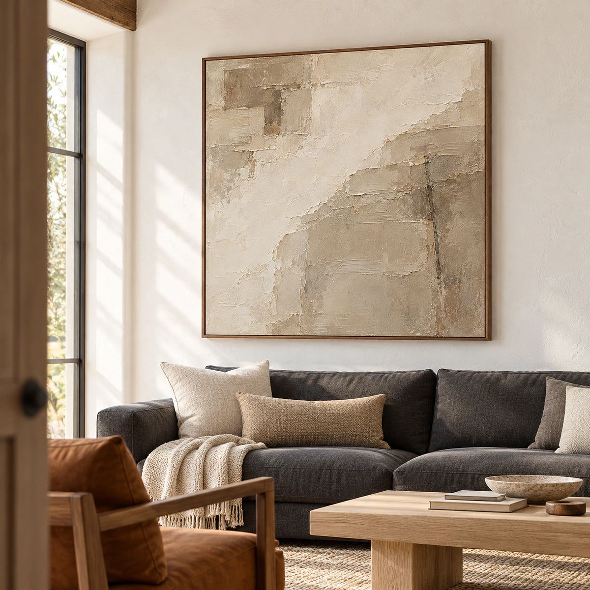 Large square ivory and taupe abstract artwork above a charcoal sofa in a bright living room with pale walls and daylight.
