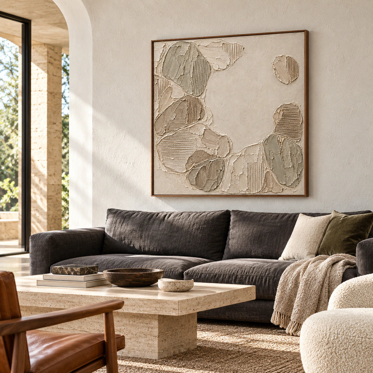 Square ivory and taupe textured painting above a charcoal sofa on a pale plaster wall, viewed from the left in a bright living room.
