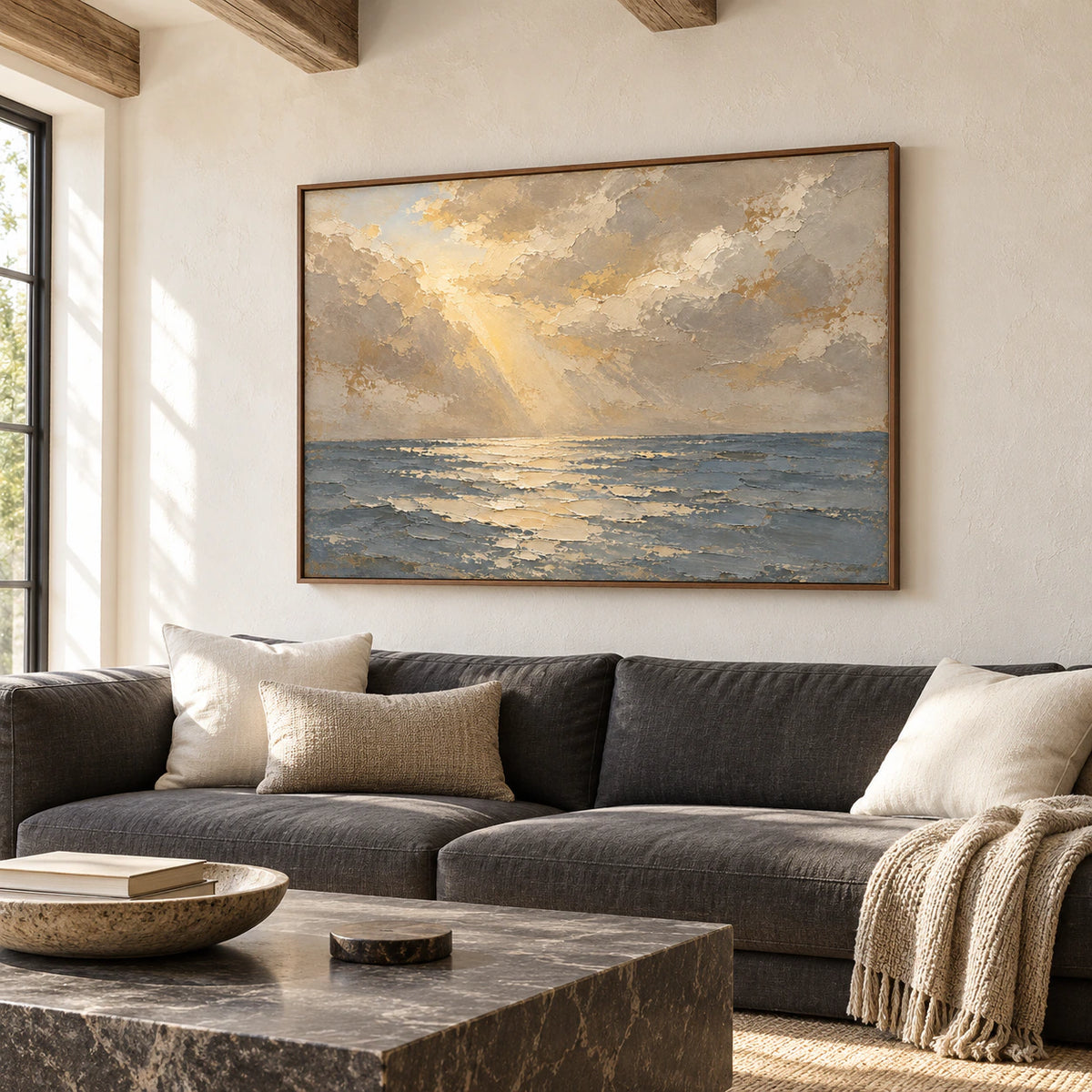 Seascape oil painting above a low charcoal sofa, viewed from the left in a bright living room with pale plaster walls and a dark stone coffee table.
