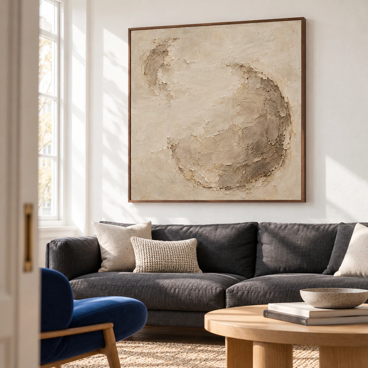 Large square warm ivory and taupe textured artwork above a charcoal sofa in a bright living room.