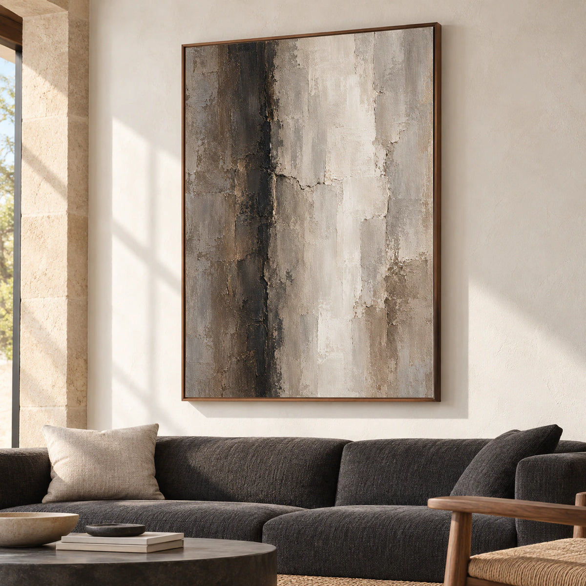 Large vertical abstract artwork above a low charcoal sofa in a bright living room with pale walls and daylight.