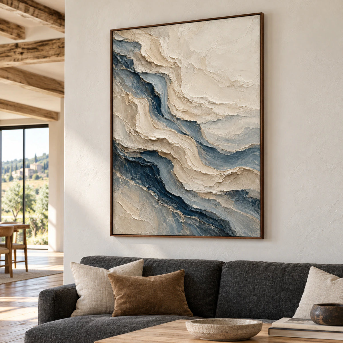Large vertical ivory and blue-gray painting above a low charcoal sofa in a bright living room with pale plaster walls and a light oak table.