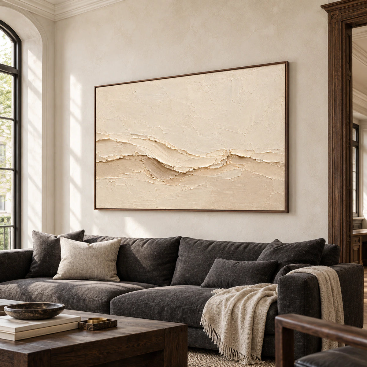 Warm ivory textured painting above a charcoal sofa against pale stone walls in a bright living room, viewed from the left at seated eye level.
