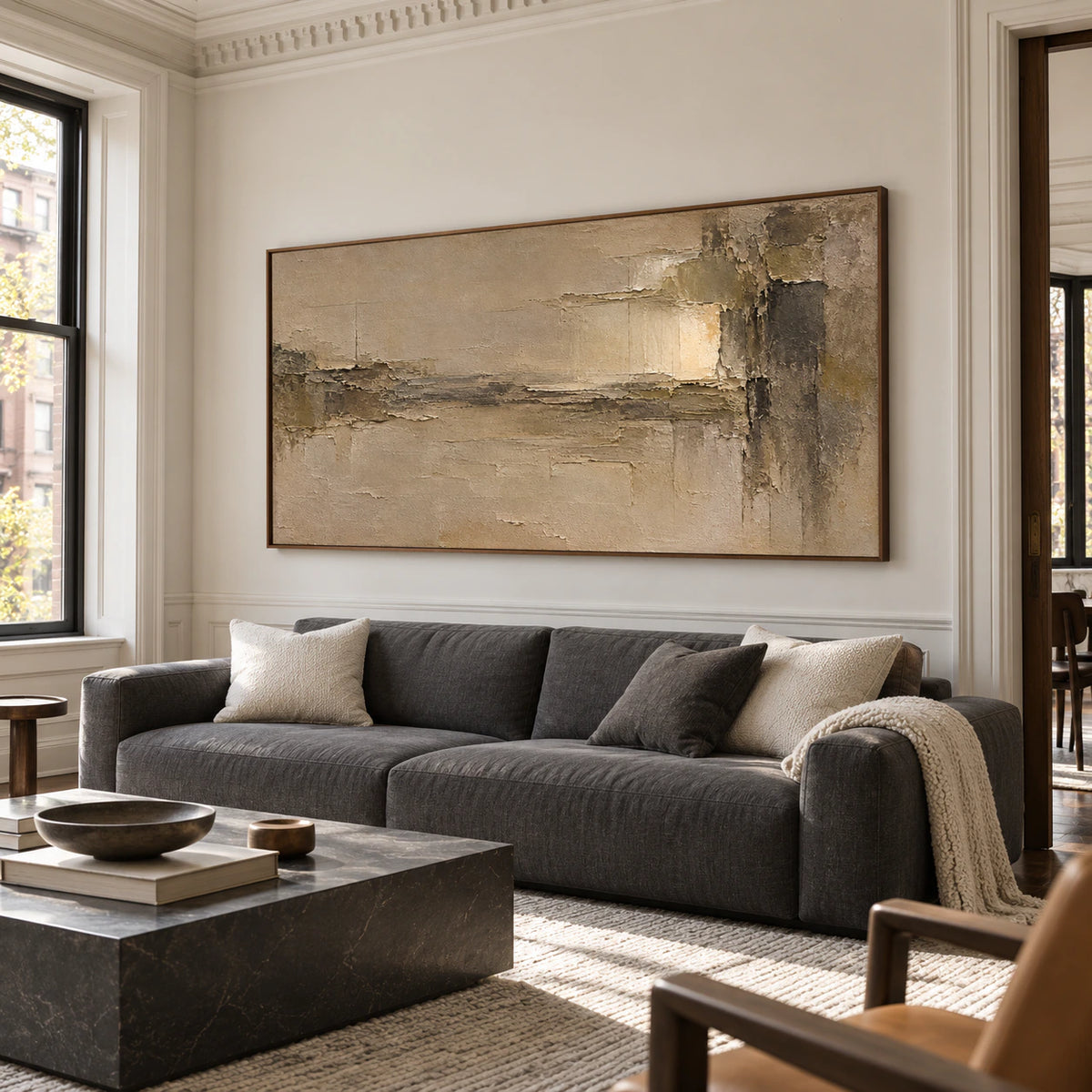 Textured ochre and umber painting above a low slate-colored sofa against a cool neutral wall in a bright living room, viewed from a right-oblique angle.