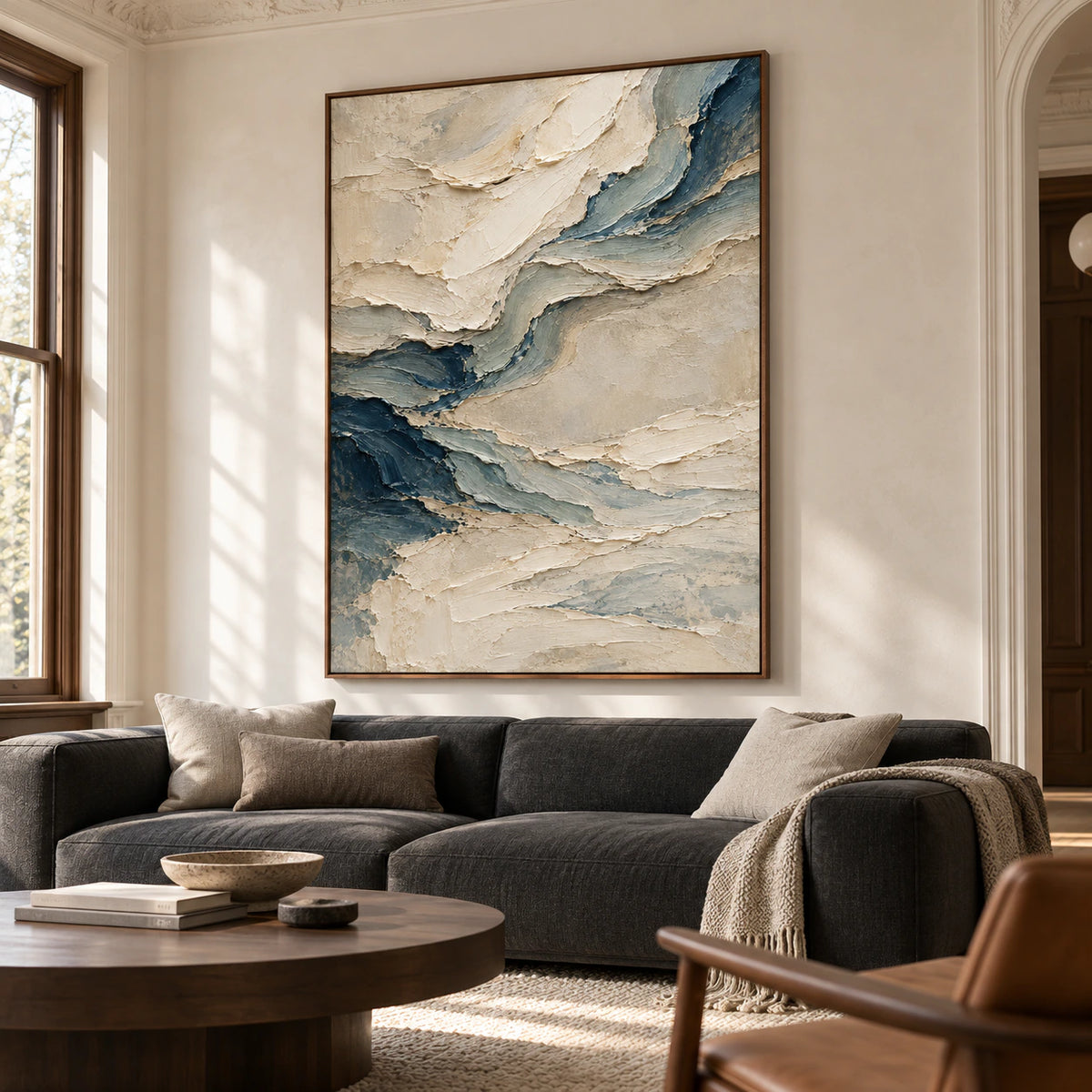 Pale-Undertow above a low charcoal sofa, viewed from the left in a bright living room with pale plaster walls and dark wood furniture.