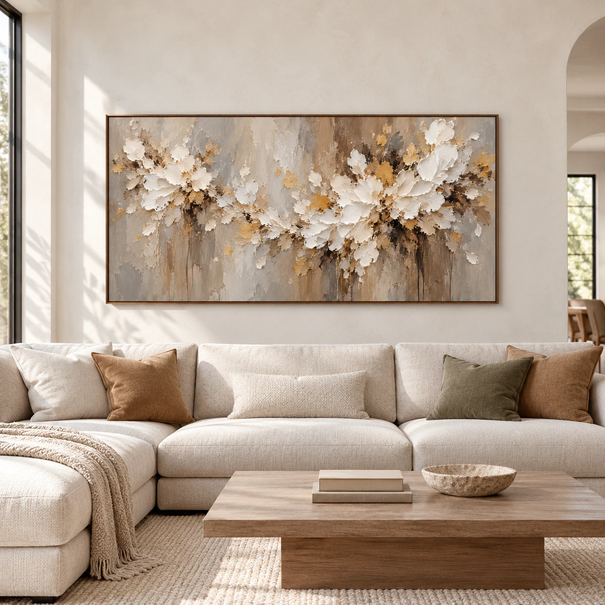 Straight-on view of a large horizontal ivory floral painting centered on a pale plaster wall in a spacious room with sparse furnishings and bright daylight.