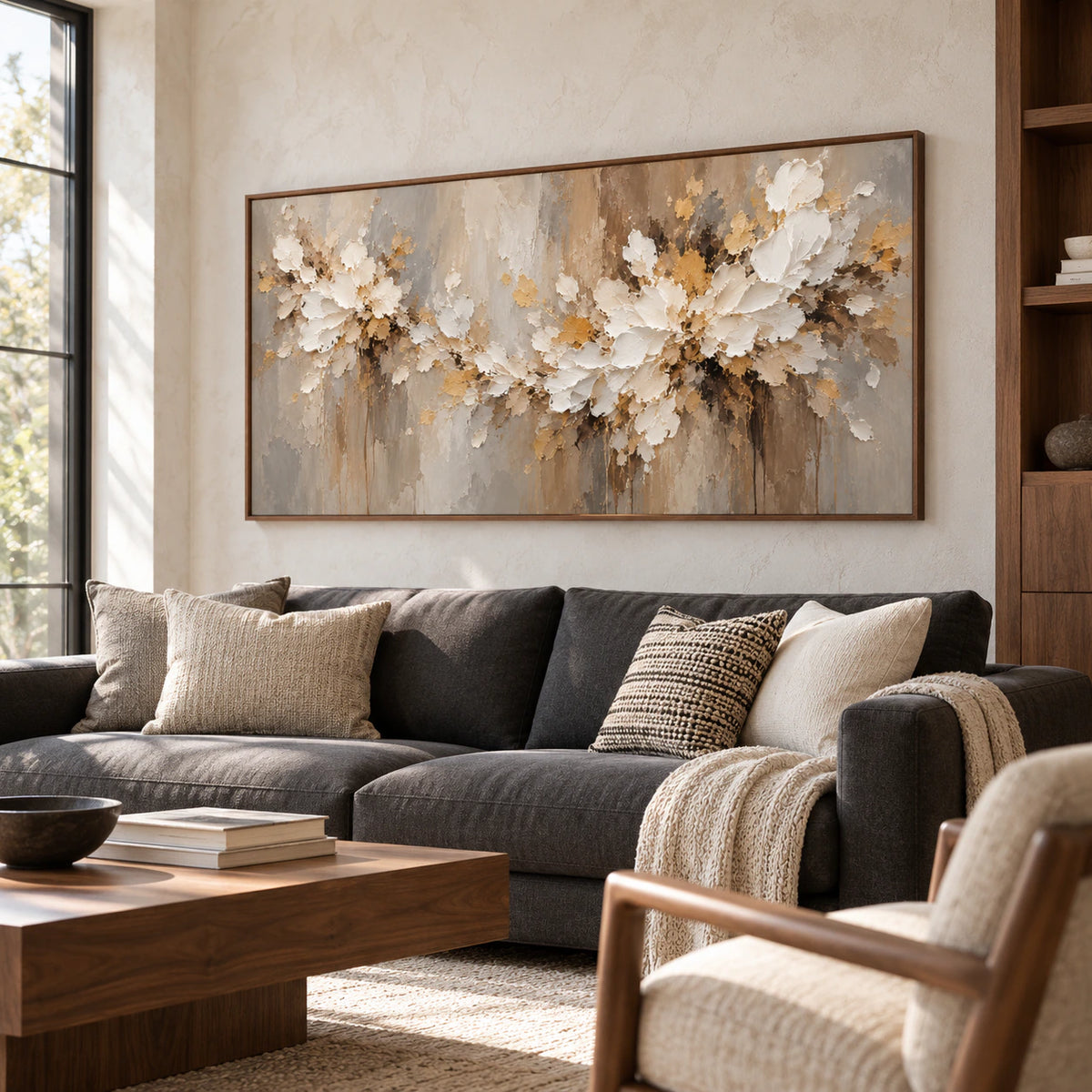 Left oblique view of a large horizontal ivory floral painting above a charcoal sofa, with a pale stone wall, dark wood accents and natural window light.