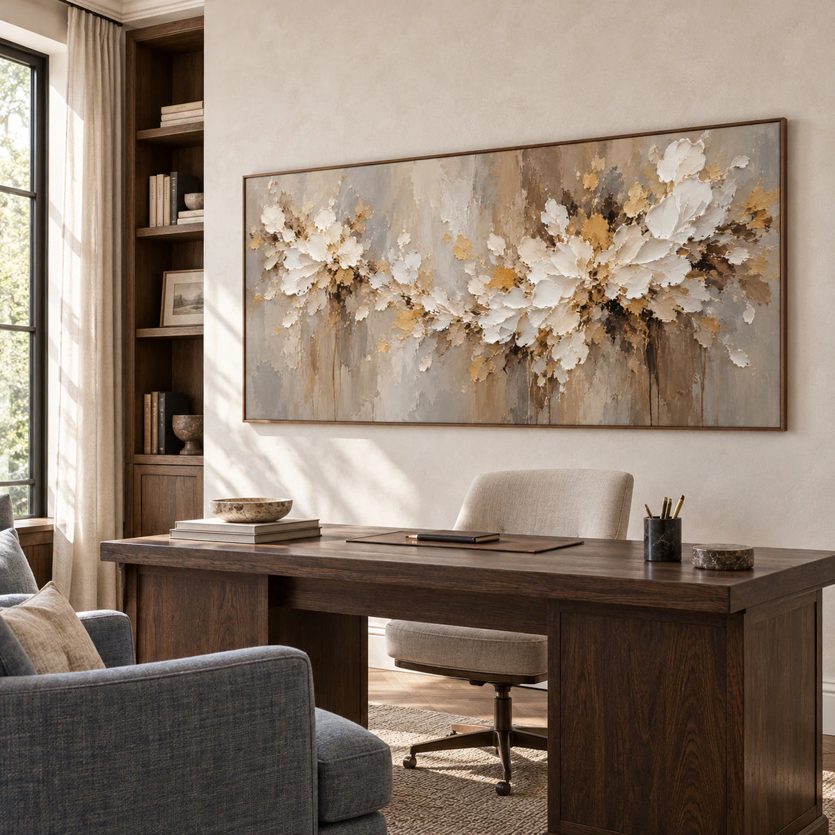 Right oblique view of a horizontal ivory floral painting above a dark writing desk, with open shelving, a blue-gray reading chair and daylight from the side.