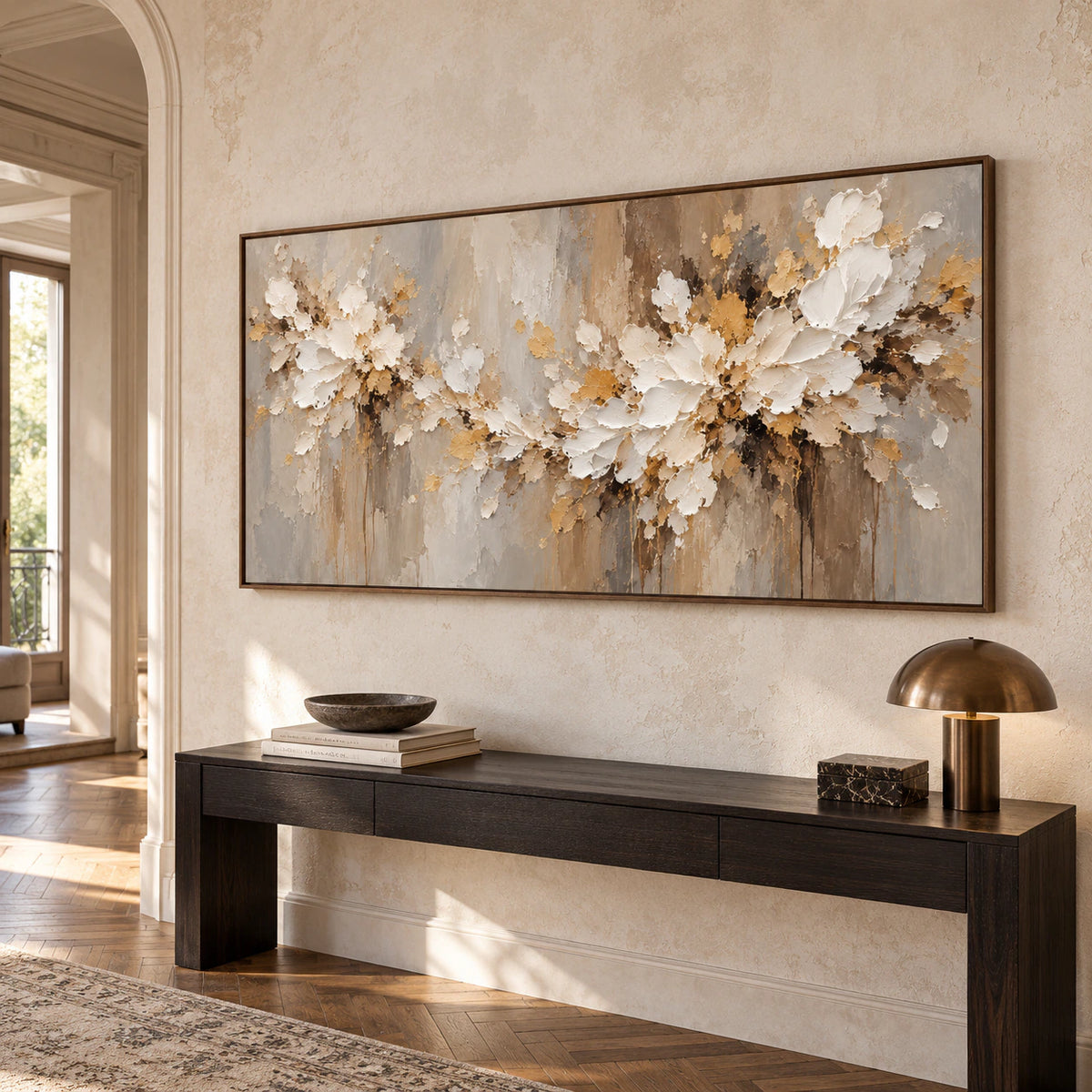 Left oblique view of a large horizontal ivory floral painting above a narrow dark console in a bright foyer with a pale limestone wall, wood parquet flooring and a tall opening.