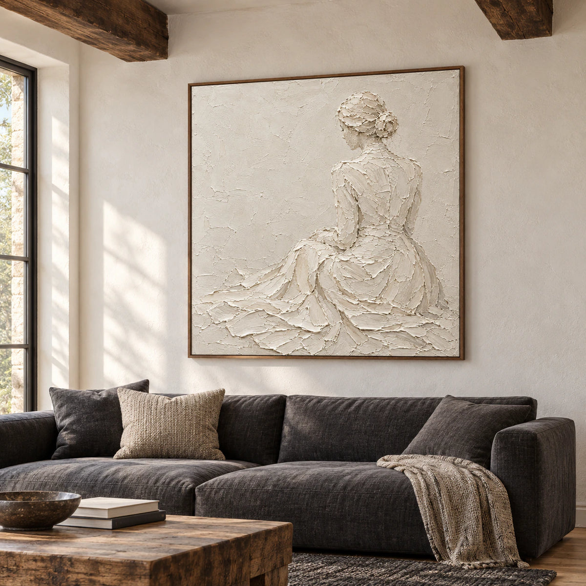 Large square ivory figure artwork above a low charcoal sofa in a bright living room with pale plaster walls and daylight from the left.