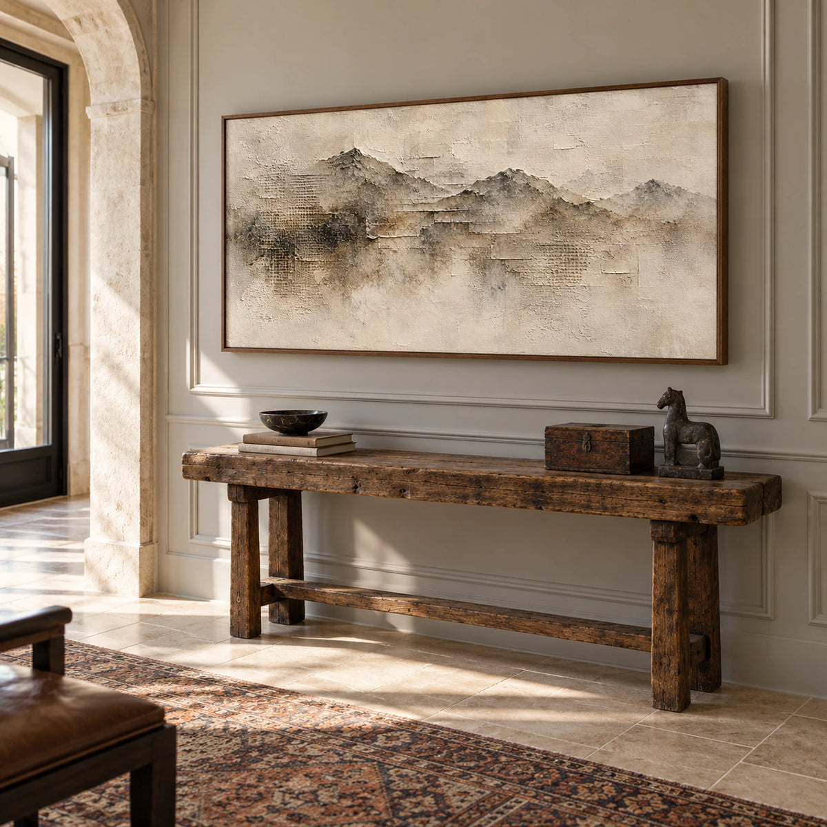 Large panoramic ridgeline painting above a rustic wooden console in a spacious foyer with tall pale stone architecture, viewed from the right in bright daylight.