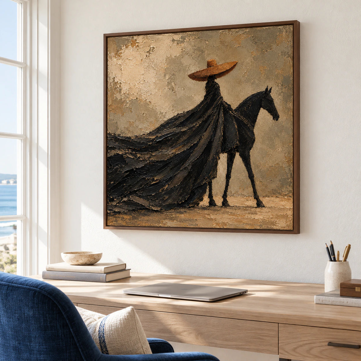 Square rider-and-horse painting on a pale wall in a bright home office with a writing desk and deep blue reading chair, viewed from a low right-oblique angle.