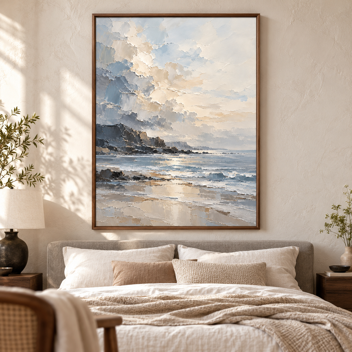 Vertical coastal painting above a low stone-gray upholstered bed with white linen, a pale wall and dark bedside accents, viewed from the left in morning light.