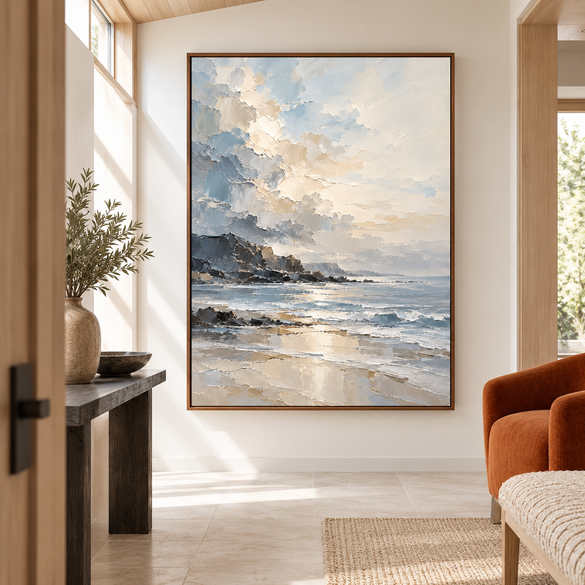 Vertical coastal painting on the far wall of a tall foyer with pale stone flooring, a narrow dark console and an ivory sculptural bench, viewed from the left.