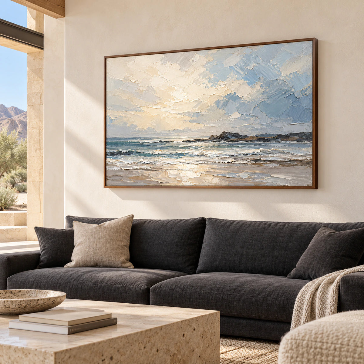 Large seascape painting above a charcoal sofa against pale plaster walls, viewed from a slight left angle at seated height in daylight.