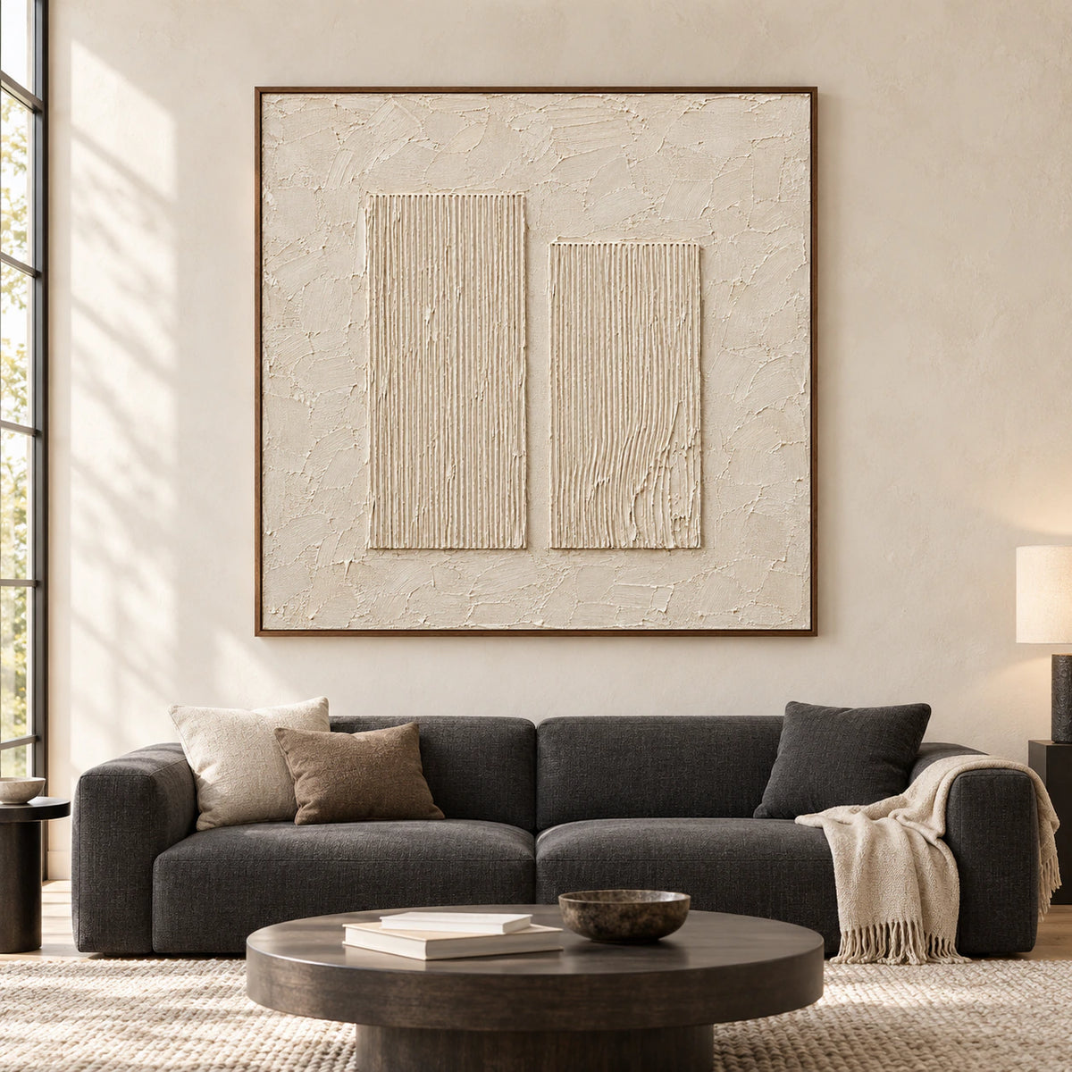 Straight-on view of a large square ivory artwork with twin ribbed forms on a pale wall in a bright living room with a low charcoal sofa.