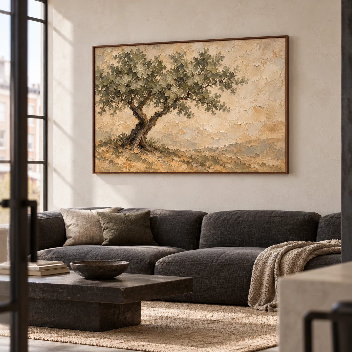 Horizontal tree painting on a pale plaster wall above a low charcoal sofa, viewed from the left with a dark coffee table in daylight.