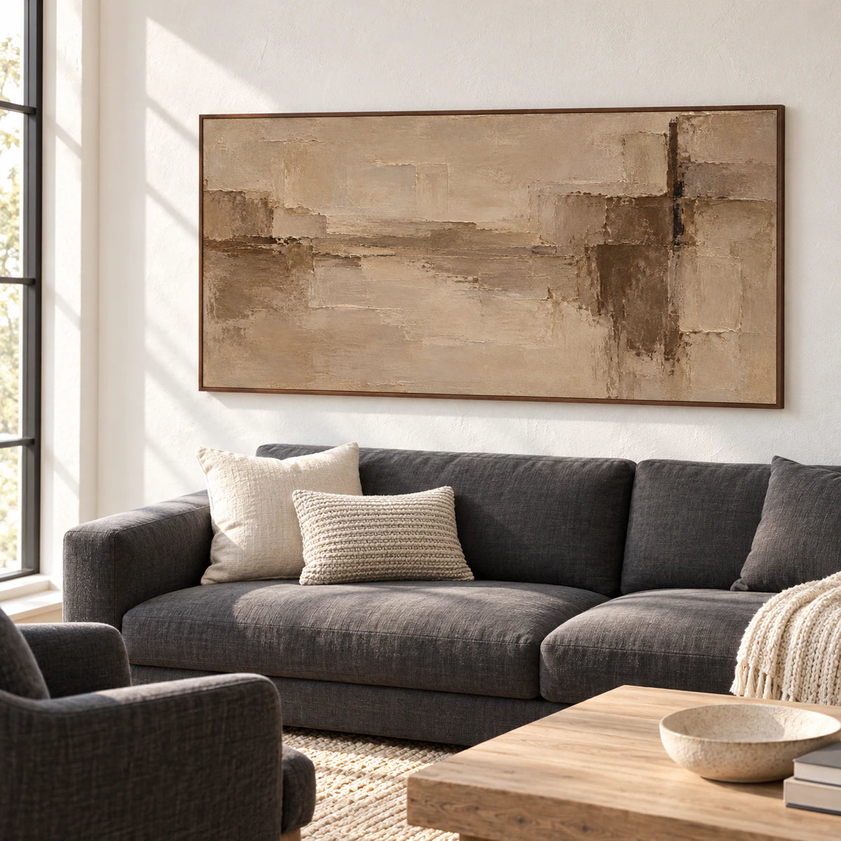 Wide sand, taupe and umber abstract painting above a charcoal sofa in a bright living room with pale plaster walls, viewed from the left.