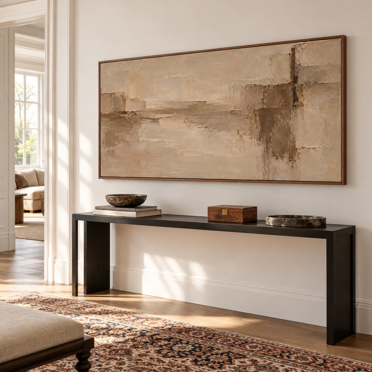 Wide sand, taupe and umber abstract painting above a slim black console in a bright foyer with pale walls, viewed from a low left angle.