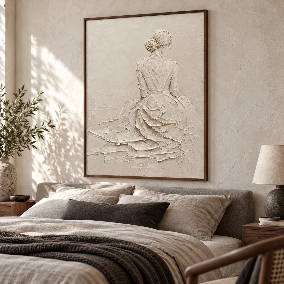 Left oblique view from near the foot of a low bed with muted gray linen and dark accents, with a large ivory figure painting above it in soft daylight.