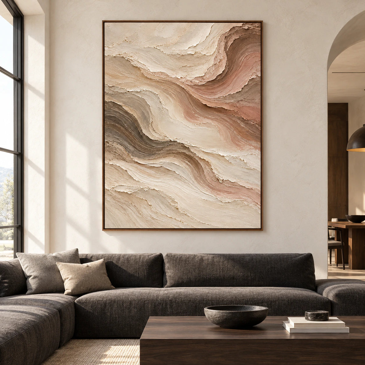 Large vertical abstract painting with flowing warm-toned ridges on a light plaster wall above a low charcoal sofa in a bright living room, viewed straight on.
